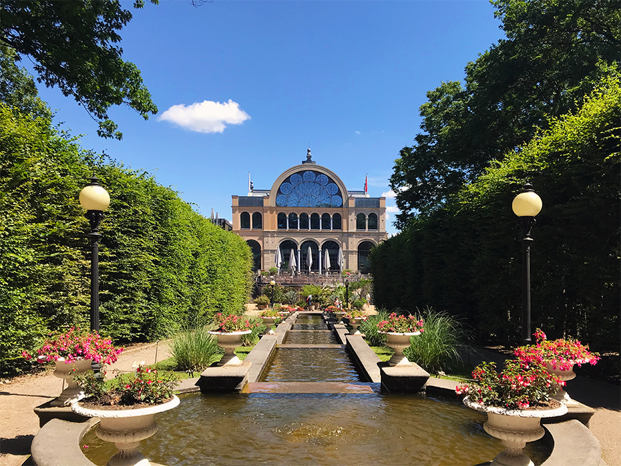 Blick über die Wassertreppe auf die Seitenfassade der Flora mit den Sonnenschirmen des Café "Dank Augusta".